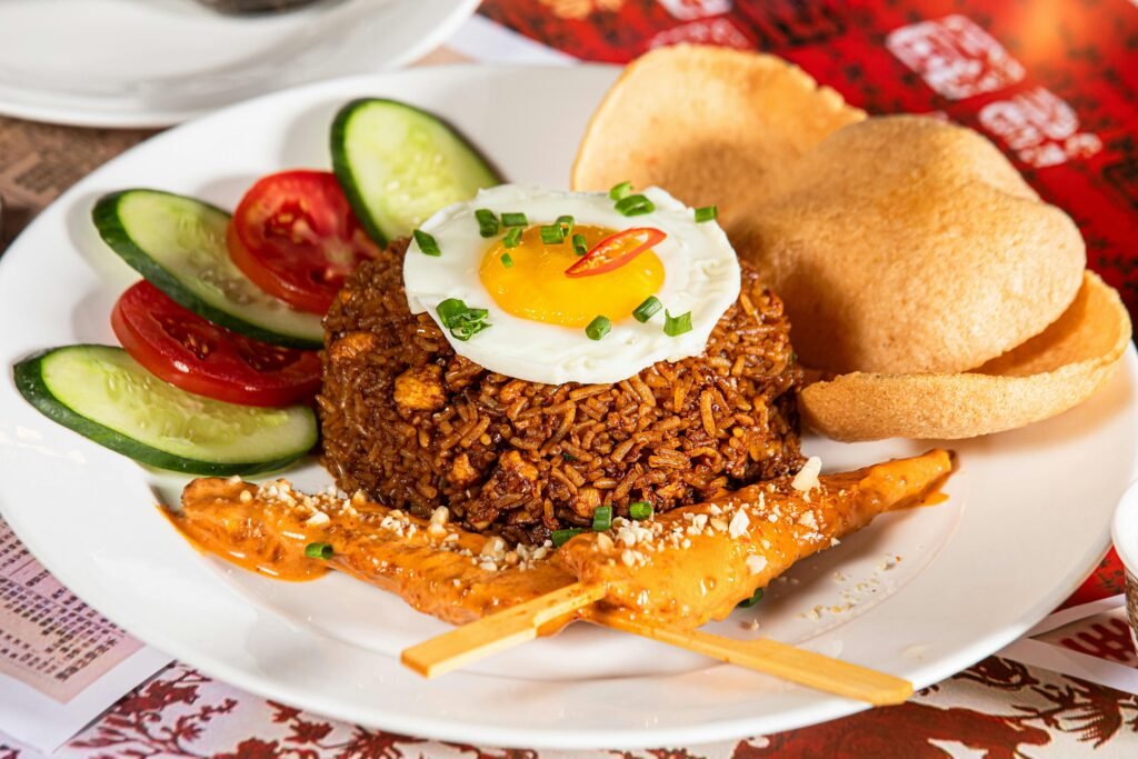A vibrant dish featuring Nasi Goreng with satay, fried egg, crackers, and fresh vegetables on a white plate.