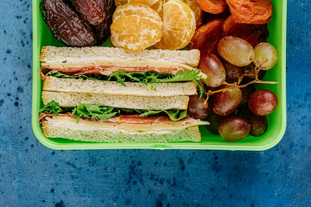 Colorful lunchbox containing ham sandwich, fresh grapes, peeled oranges, and dates on a blue background.